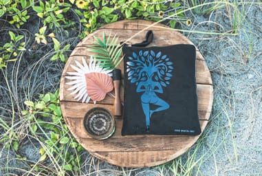 Good Mantra branded tote bag with tree pose design displayed on wooden table with plants.