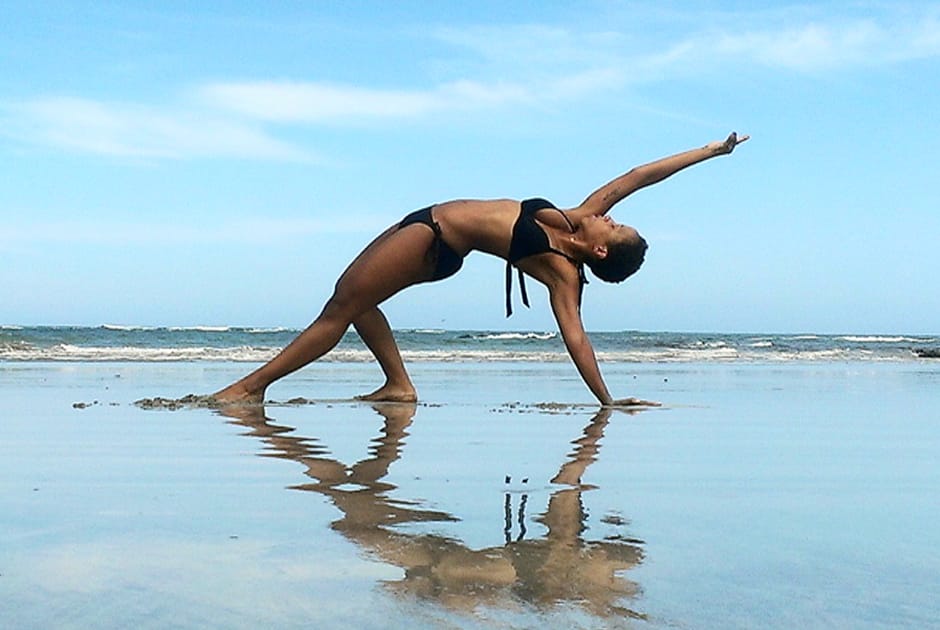 Nicole Spence performing extended triangle pose on wet sand beach with water reflection.
