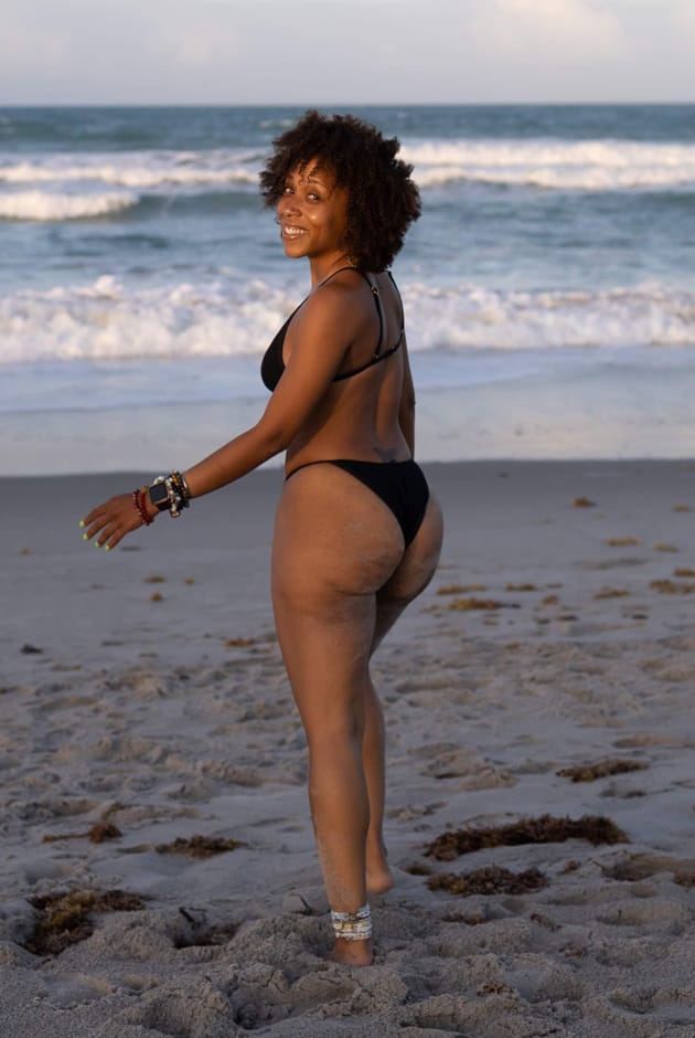Nicole Spence smiling at camera on beach in black bikini celebrating body positivity and wellness.