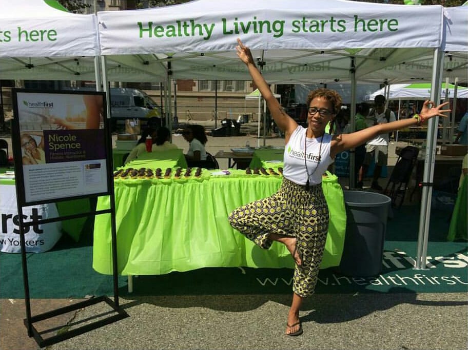 Nicole Spence in tree pose at outdoor health expo booth with healthy living starts here banner.